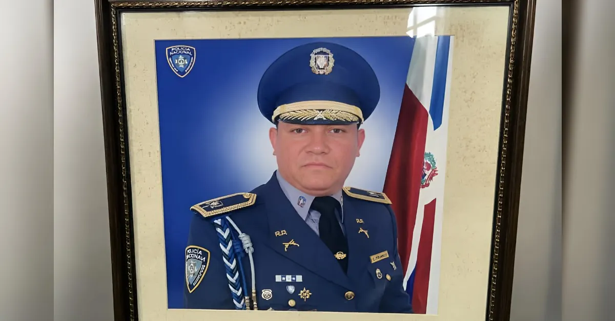 Framed portrait of a Dominican police officer in ceremonial uniform with medals, blue background and flag beside him.