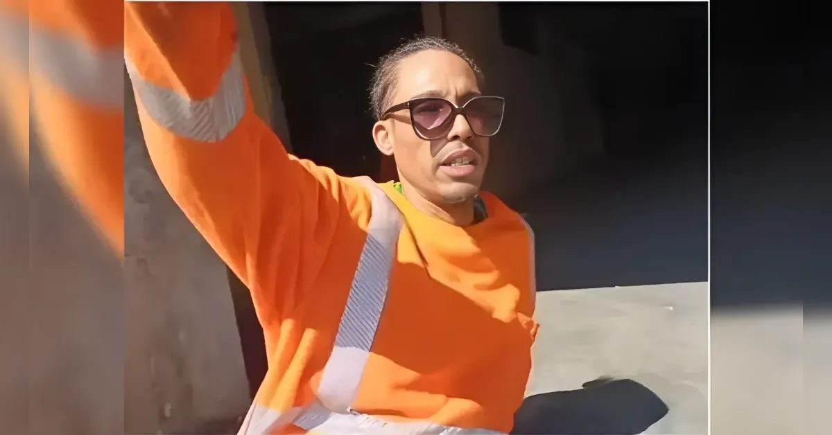 Construction worker in an orange safety suit and sunglasses raises an arm toward the camera near a dark tunnel entrance.
