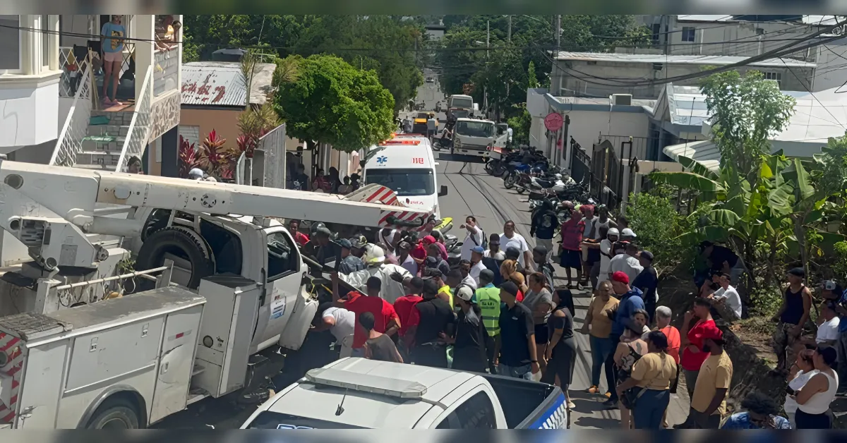 Crowd of people on a sunny urban street gathered around a large white bucket truck and an ambulance in the background, likely at an emergency scene.
