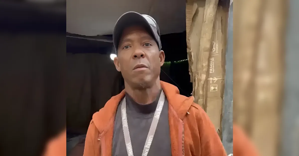 Man wearing a gray cap and orange jacket stands in a dim warehouse doorway, with a lanyard around his neck and cardboard boxes in the background.