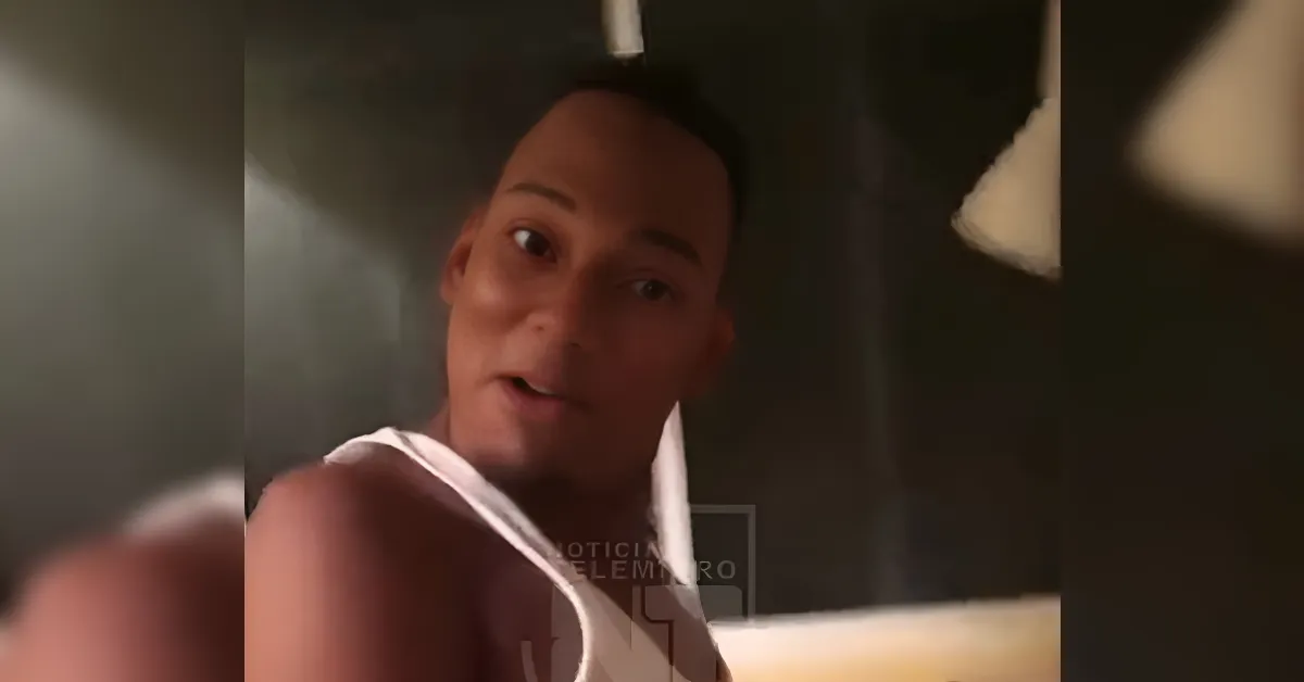 Close-up of a man in a white sleeveless shirt looking at the camera with a surprised expression in a dim indoor space.