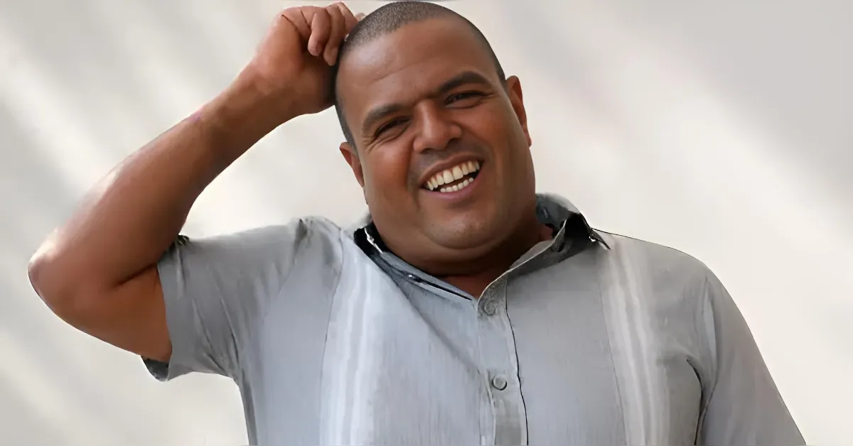 Smiling man in a light gray button-down shirt, hand resting on top of his head.