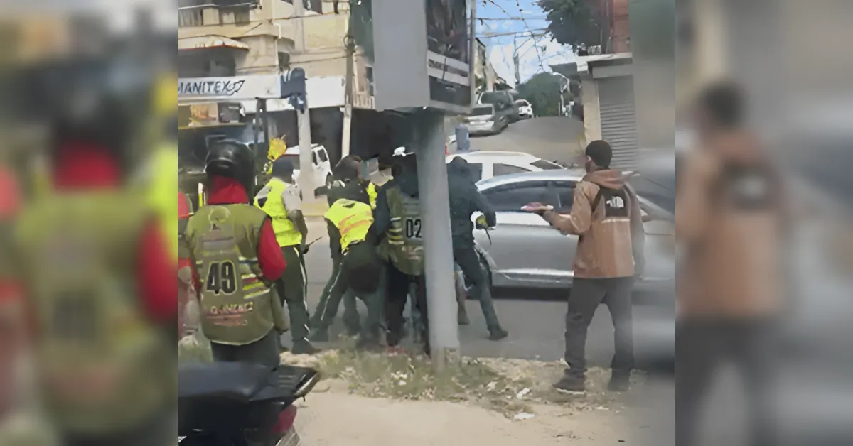People wearing high-visibility vests crowd around a car on a city street, some pushing or arguing near a light pole.