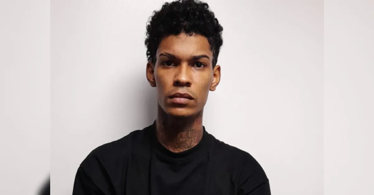 Young man with short curly hair and neck tattoos wearing a black shirt, looking straight at the camera against a light background.