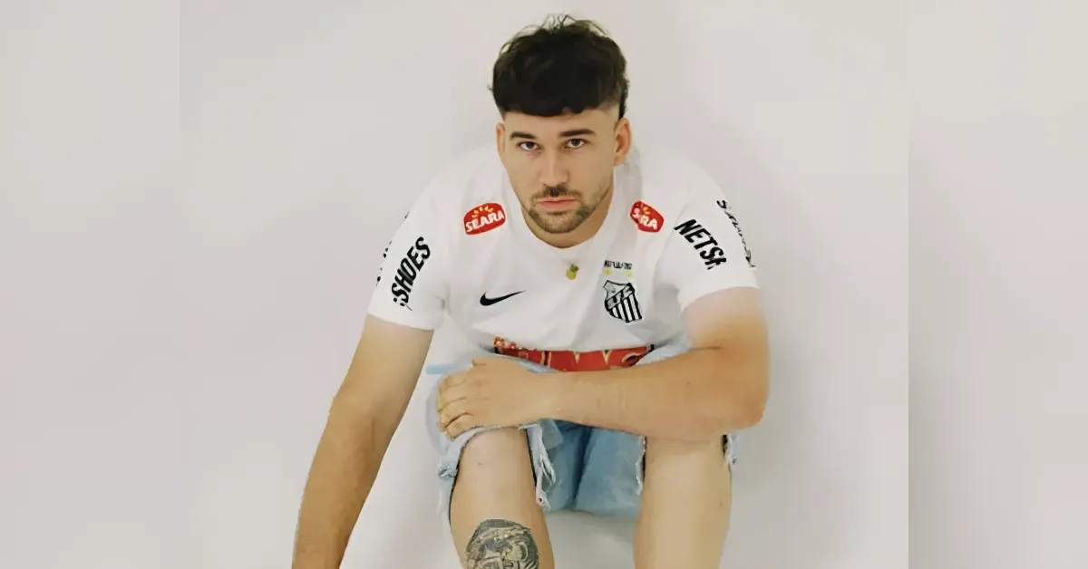 Soccer player in a white team jersey sitting on the floor, looking at the camera with a tattoo on his leg.
