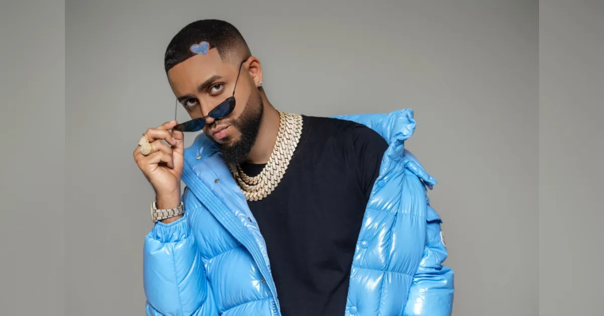 Portrait of a man in a bright blue puffer jacket, black tee, and sunglasses, tilting his shades toward the camera with gold jewelry.
