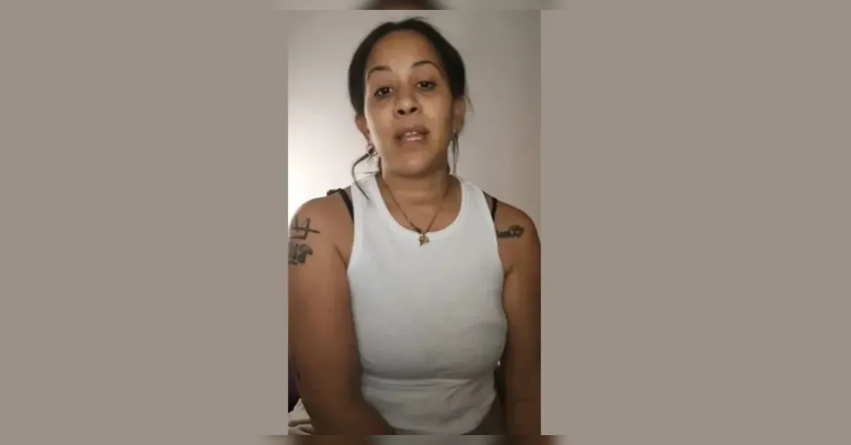 Woman in a white sleeveless top speaks directly to the camera, indoors, with visible shoulder tattoos and a necklace.