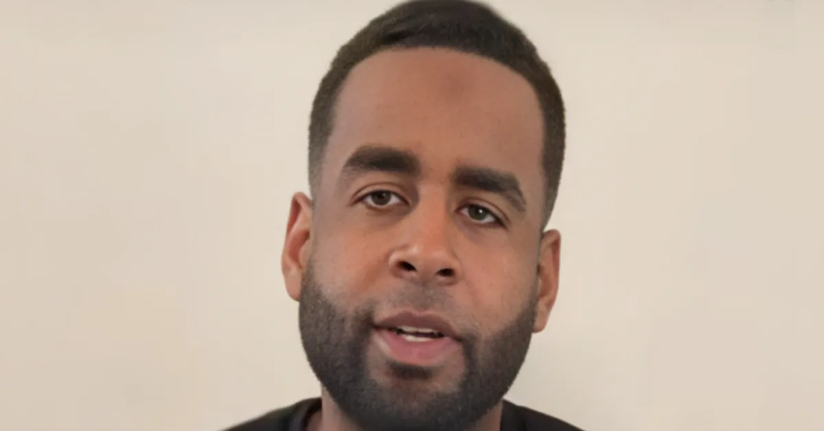 Close-up of a man with short dark hair and beard speaking directly to the camera against a plain beige background.