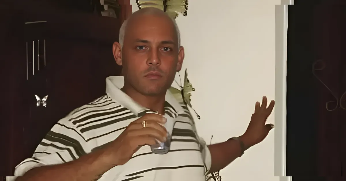 Man with a shaved head in a white-and-black striped polo holding a drink, leaning against a wall and looking at the camera.