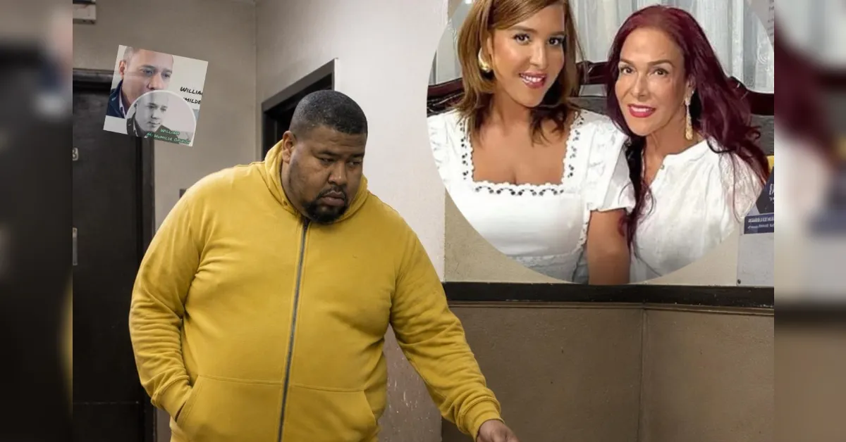 Heavyset man in a mustard yellow zippered hoodie walks through a hallway; a round poster of two smiling women in white blouses hangs on the wall behind him.