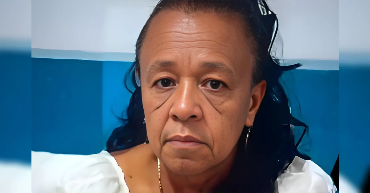 Close-up of a middle-aged woman with dark hair, wearing a white blouse, looking directly at the camera against a blue background.
