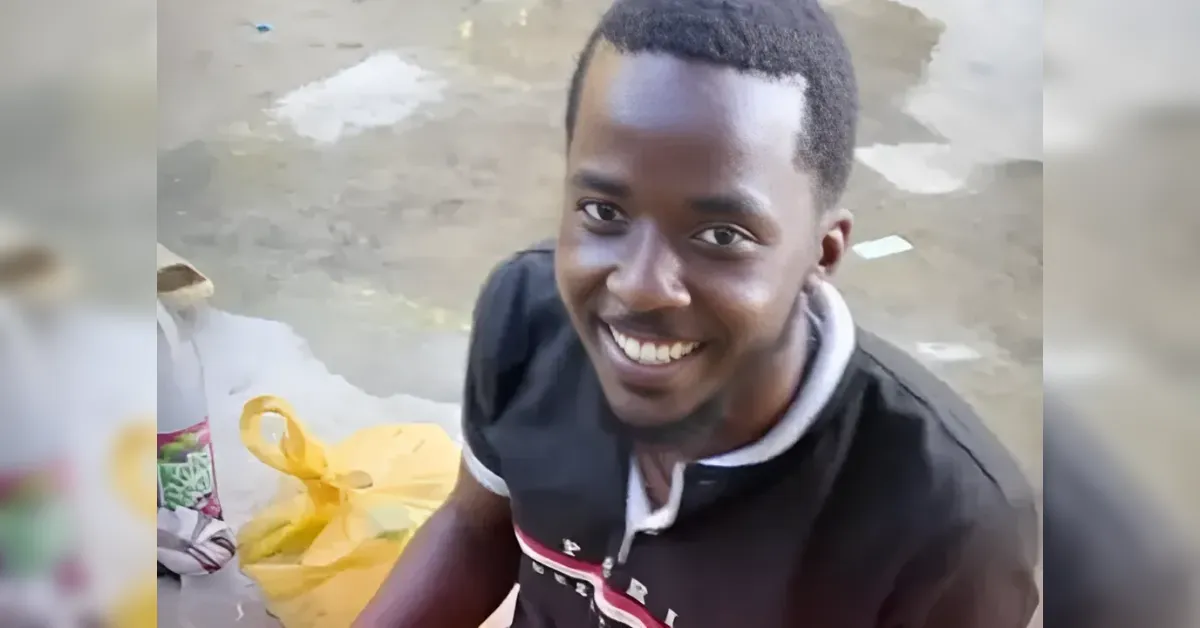 Smiling young man with short hair, wearing a black polo with white trim, outdoors near yellow bags on a wet pavement.