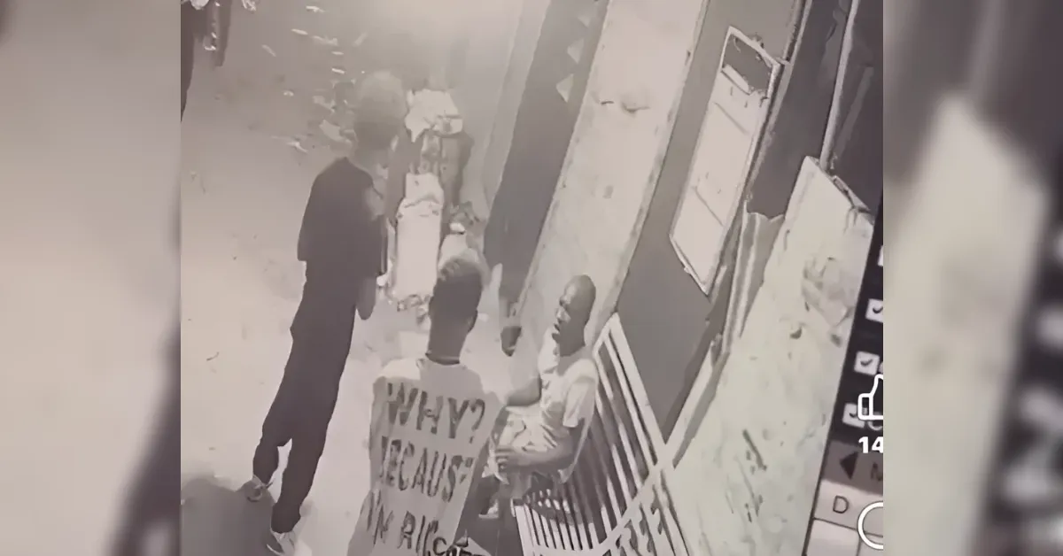 Surveillance video frame of a narrow city street: a man in dark clothing stands facing another man who sits on a bench outside a storefront; the standing man has a large message on his back (text visible but not fully readable).
