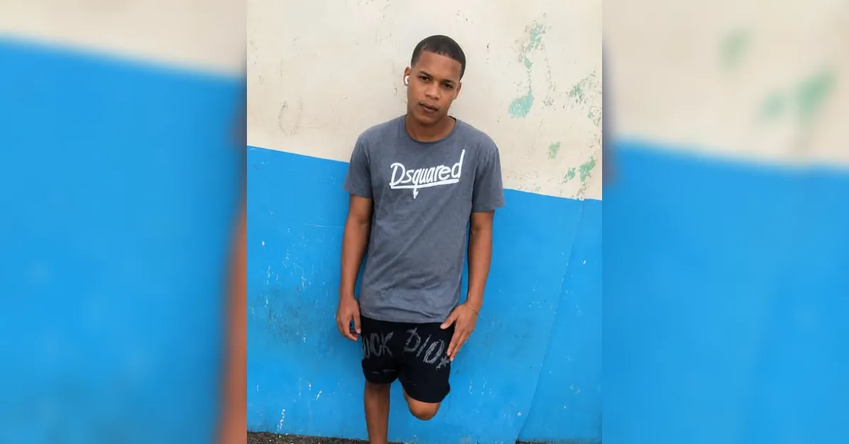 Young man leaning against a blue and white wall, wearing a gray t-shirt and black shorts.