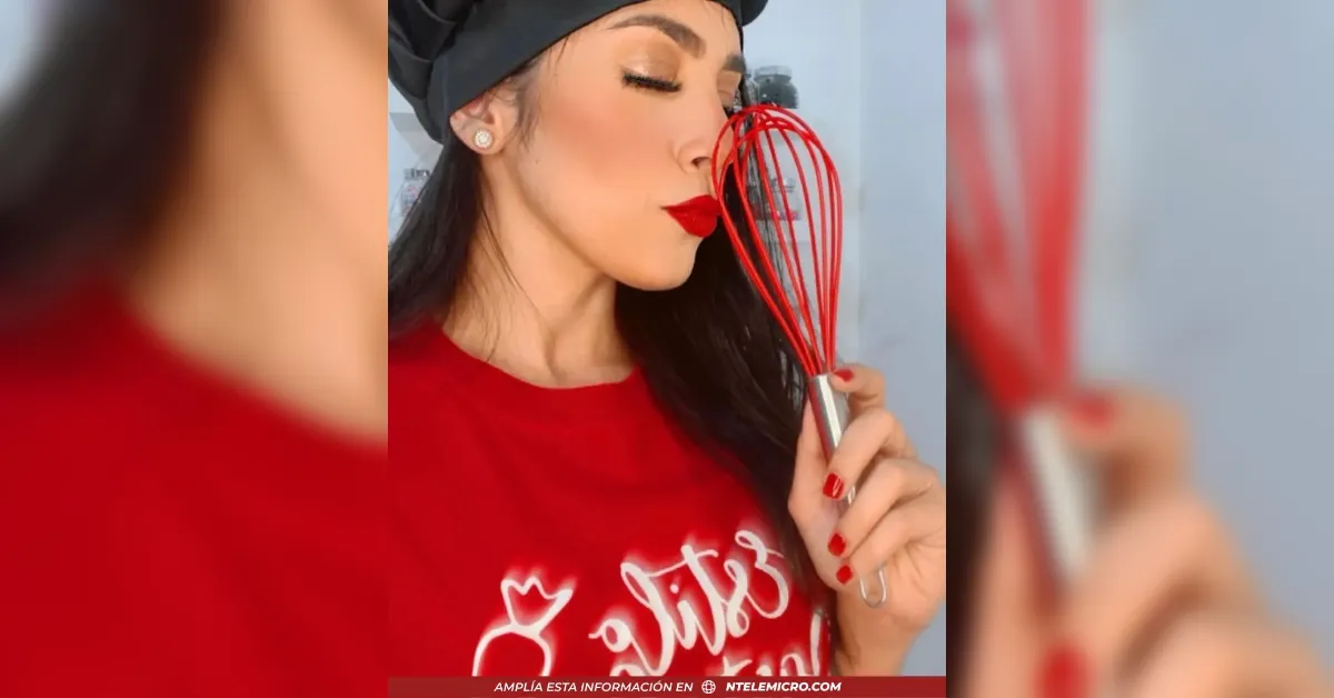 Woman in a red top and black headscarf holds a red whisk near her nose, eyes closed, bold red lipstick, posing for a playful kitchen shot.