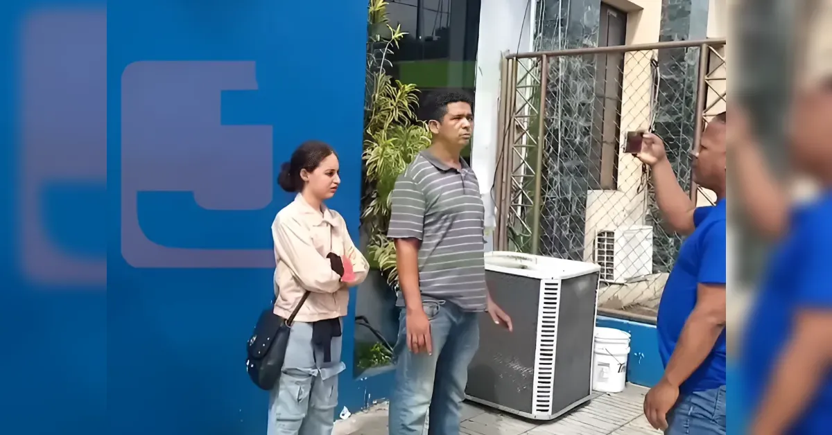 Three people outside by a blue wall: a man in a striped shirt, a woman with a beige jacket and a shoulder bag, and another man holding a phone to take a photo near an outdoor unit.