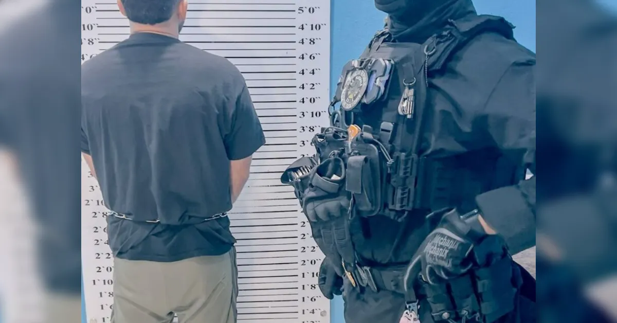 Person in a gray shirt facing a height-measurement wall as if for a mugshot, with a heavily armed officer in tactical gear nearby.