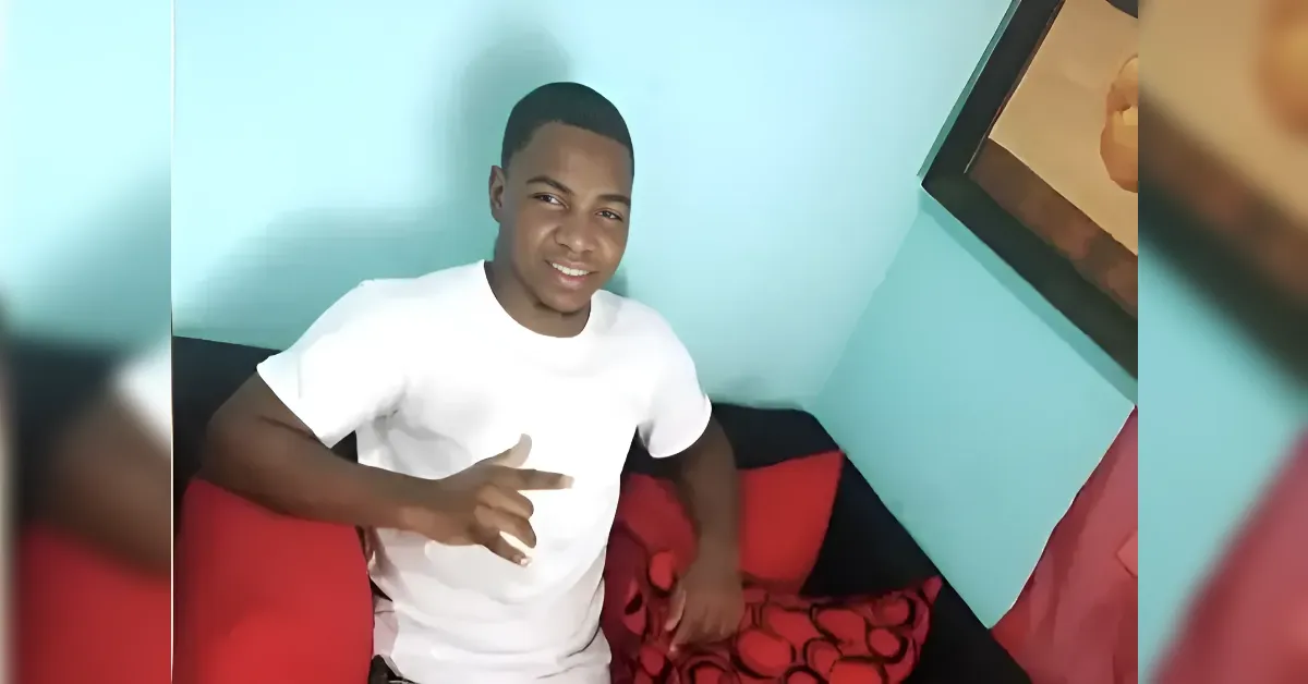 Young man in a white t-shirt sits on a red-and-black couch, smiling and making a peace sign with his fingers.