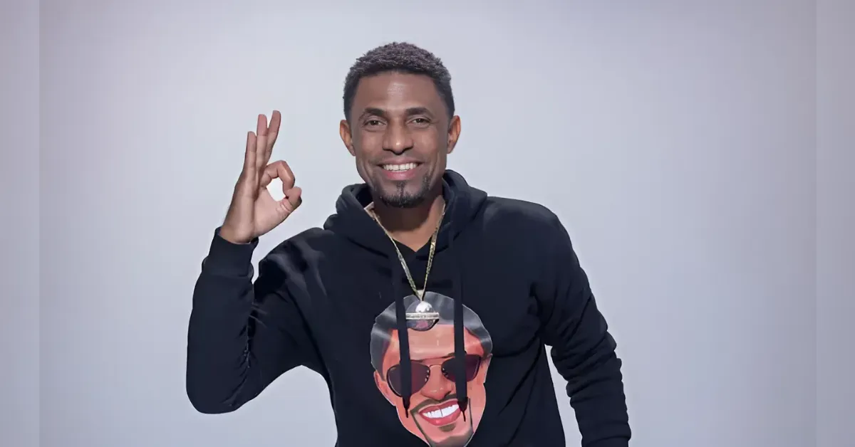 Smiling man in a black hoodie giving an OK sign with his right hand, against a light gray background.