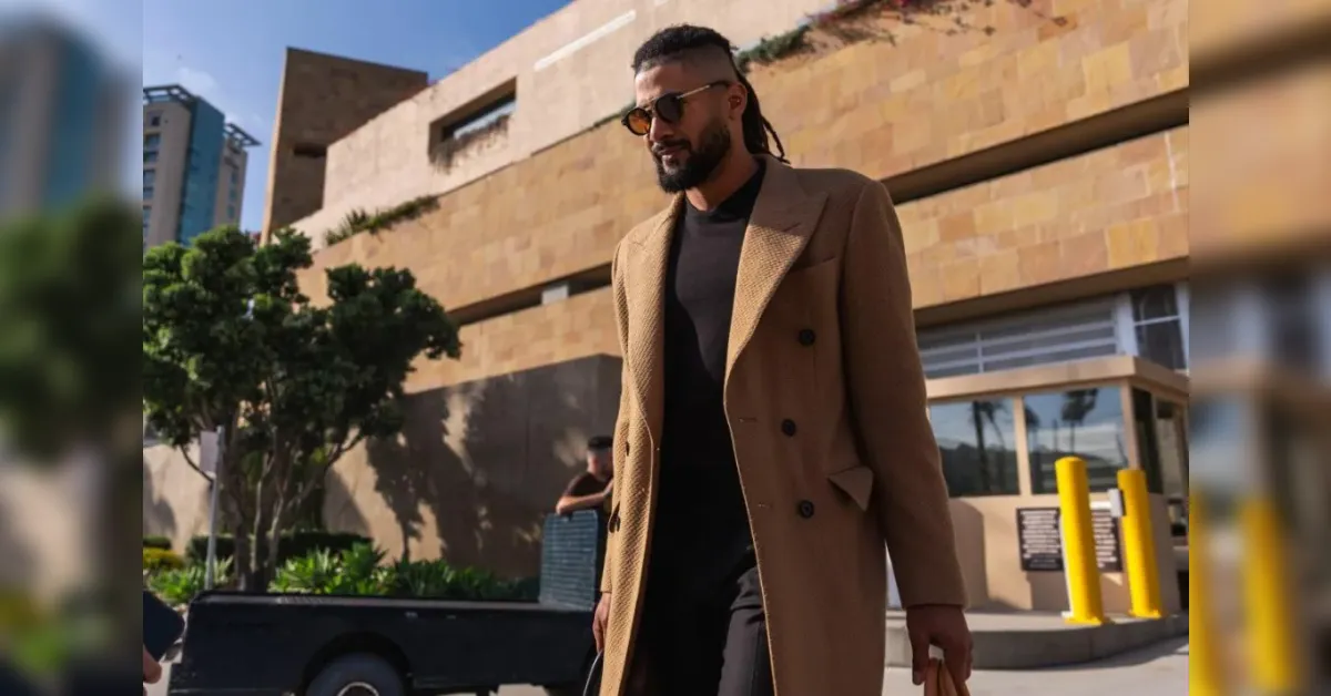 Man wearing a camel overcoat and black outfit walking outdoors near a modern beige brick building on a sunny day.