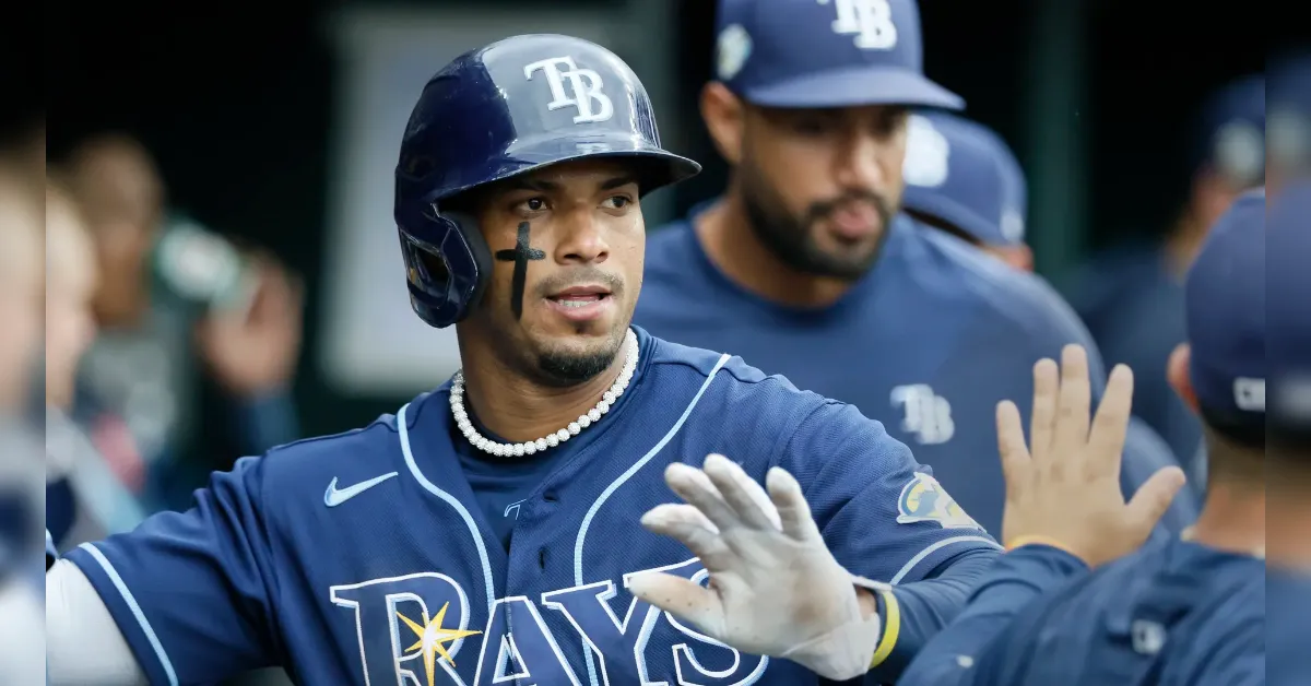 Baseball player from the Rays high-fiving a teammate in the dugout, wearing a navy uniform with TB on helmet and 'Rays' across the chest, facial paint on his cheek.