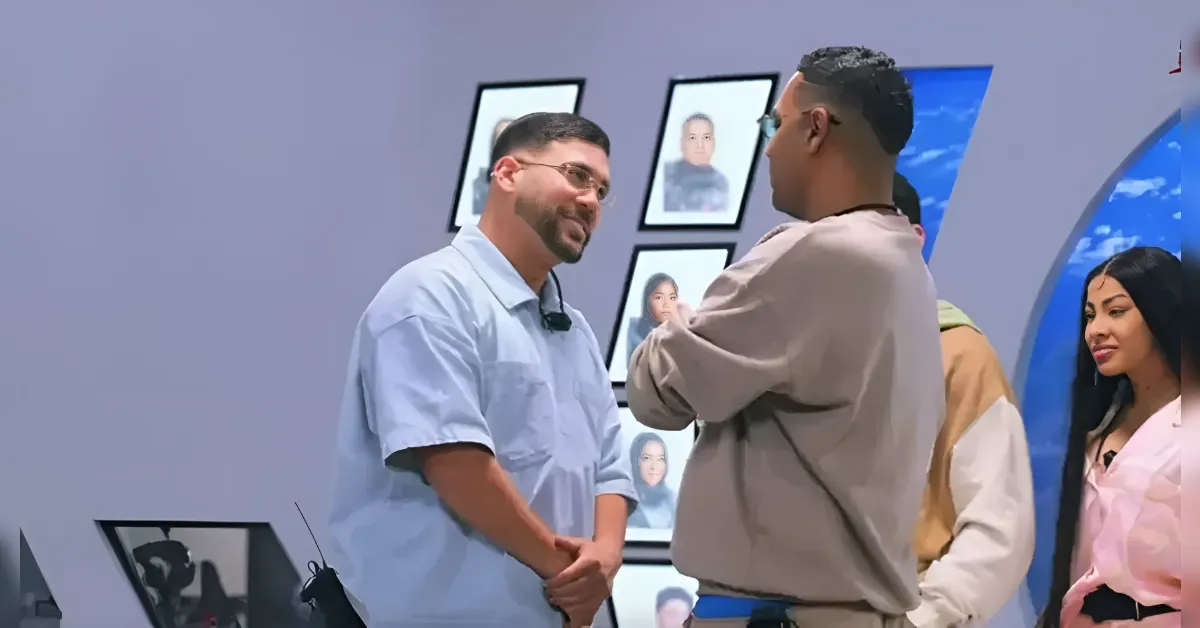 Two men in a casual conversation indoors, with framed portraits on the wall behind them.
