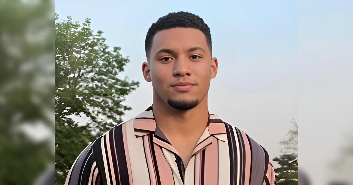 Young man standing outdoors, wearing a pink, beige, black striped shirt, looking at the camera, trees in the background.