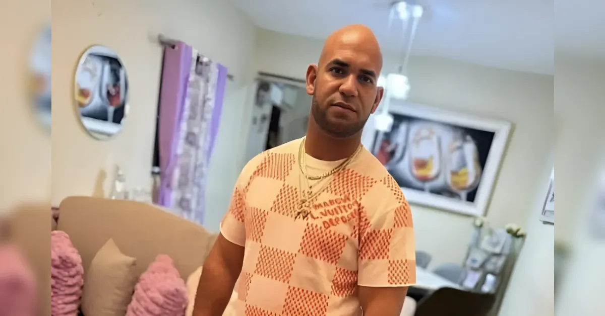 Man with a shaved head wearing a peach and orange checkered T-shirt stands in a cozy living room with pink cushions.