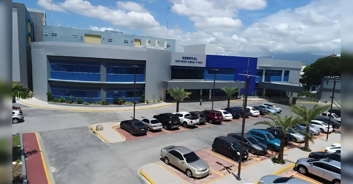 Hospital exterior with blue facade and a row of parked cars in a sunny lot