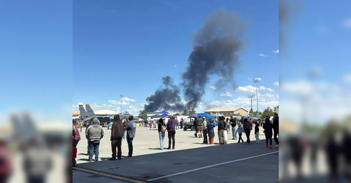 Crowd of people on an airfield watching a plane as a tall plume of black smoke rises behind them in the distance.