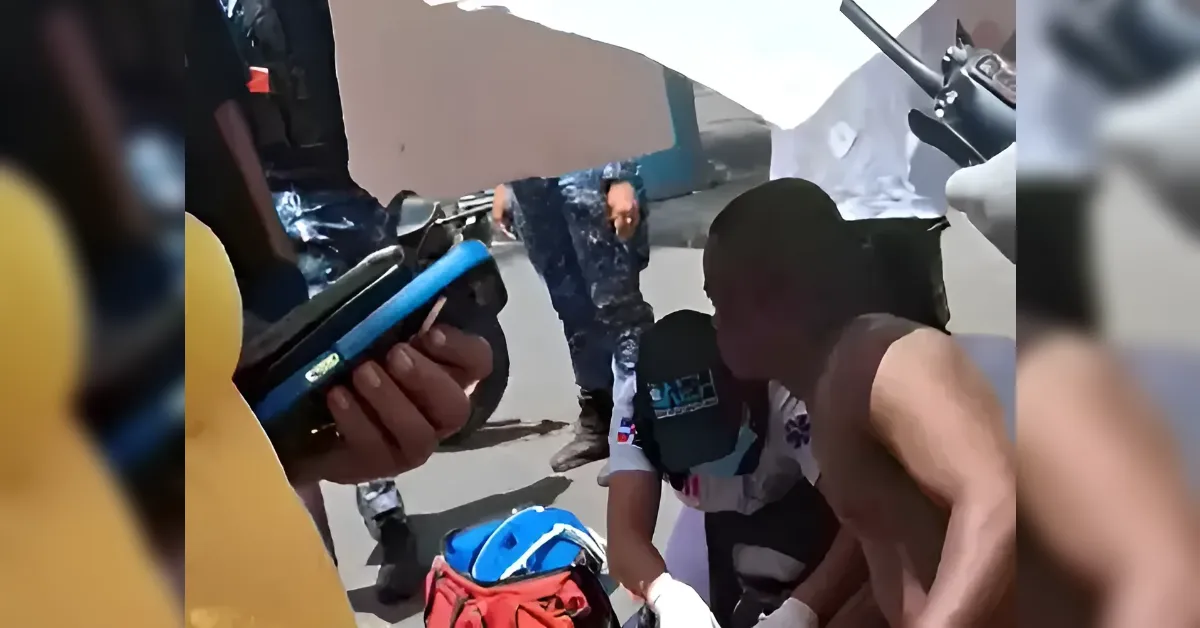 Two men crouch beside a man on the ground as others help, with a blue scooter and bags nearby.