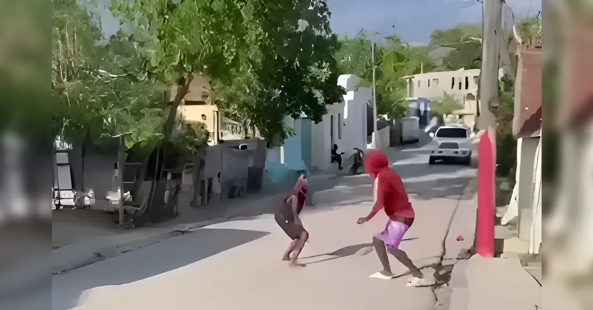 Two people in a sunny street playing or dancing, one in a red hooded sweatshirt and purple shorts, a tree-lined residential block in the background.