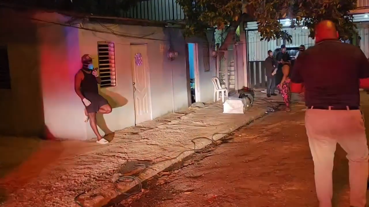 Nighttime street scene with debris along the curb, a man leaning against a wall, and people near a doorway.