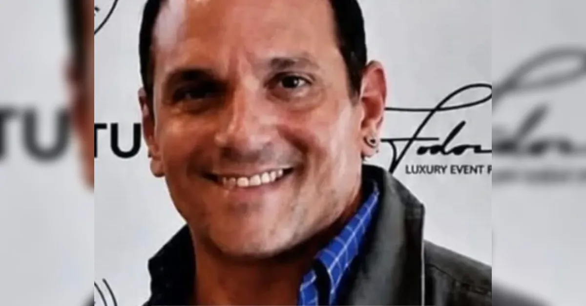 Smiling man in a blue checkered shirt and dark jacket at an event, with a backdrop featuring logos in the background