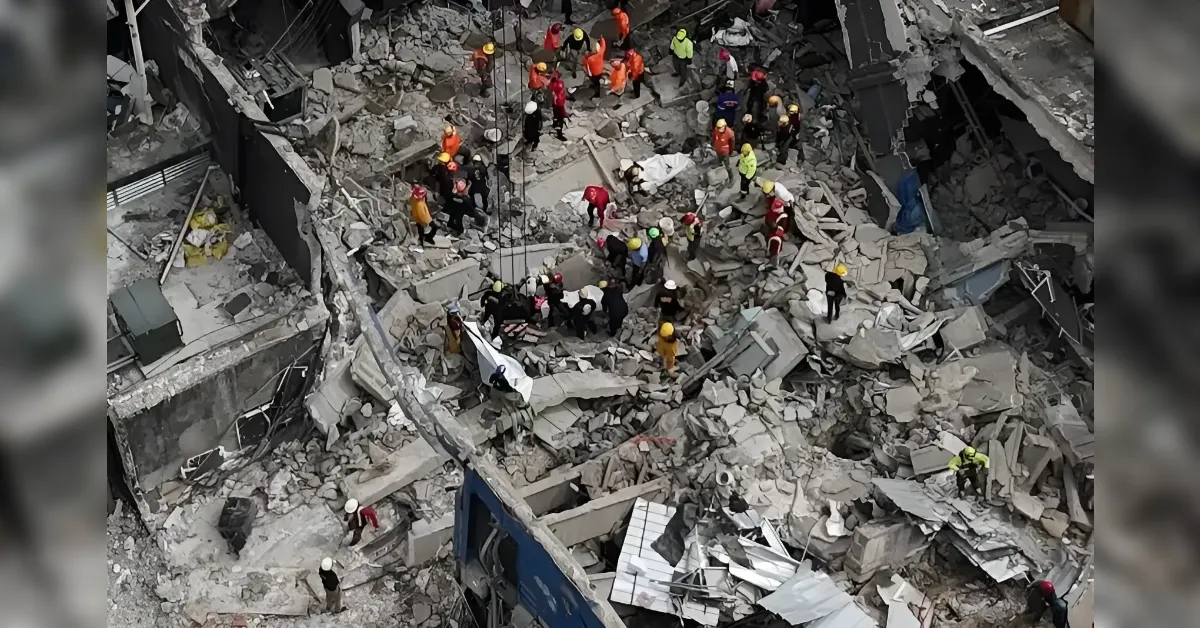 Rescue workers in hard hats search through rubble of a collapsed building, using ropes and a stretcher amid debris and rescue equipment.