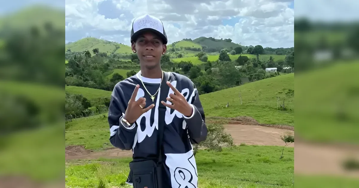 Young man in a cap and dark jacket stands on a green hillside, making hand signs toward the camera.