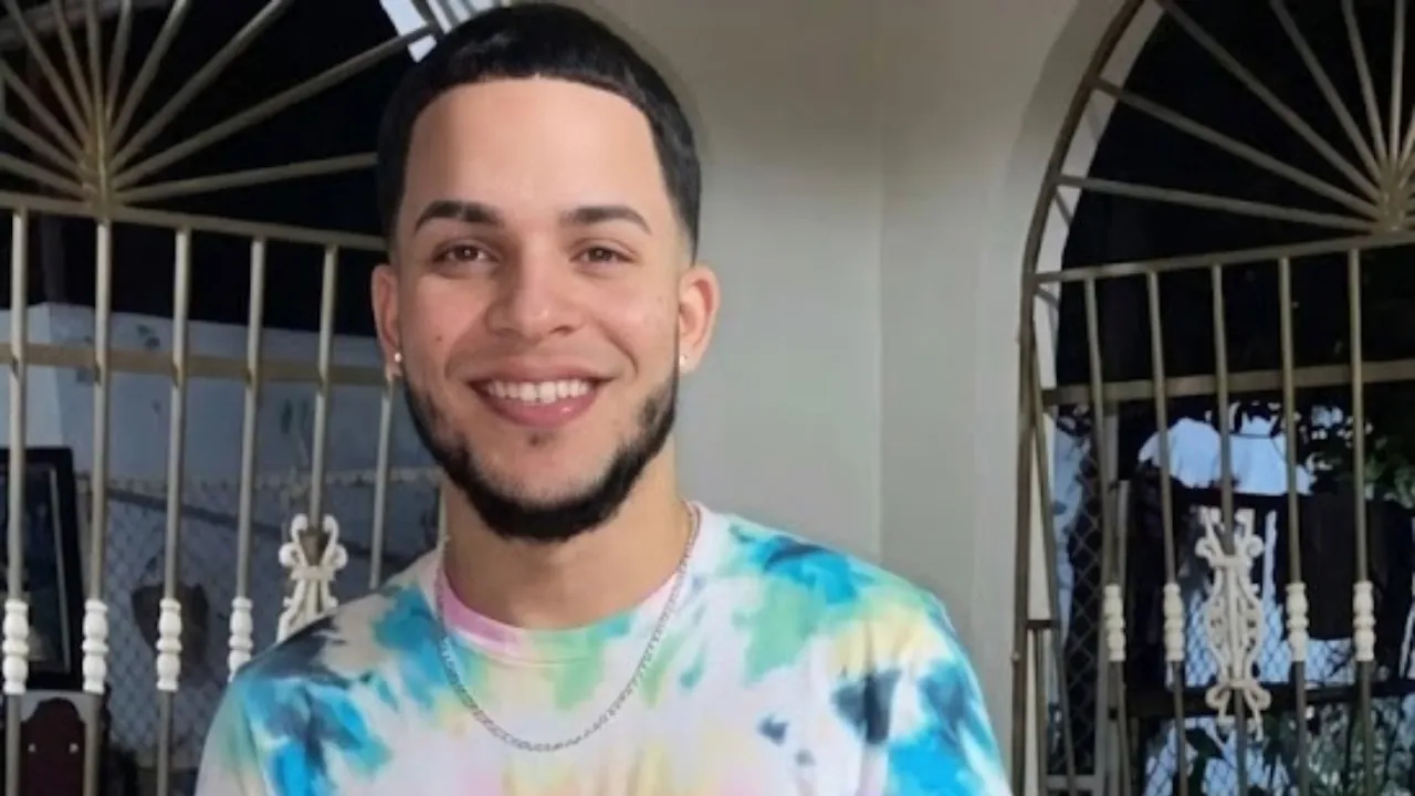 Young man smiling at the camera, wearing a pastel tie-dye shirt and silver chain, standing by a decorative gate.