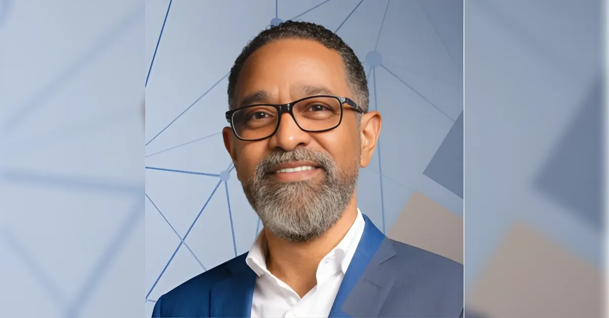 Smiling man with glasses and a gray beard in a navy suit and white shirt, posing for a head-and-shoulders portrait against a blue geometric background.