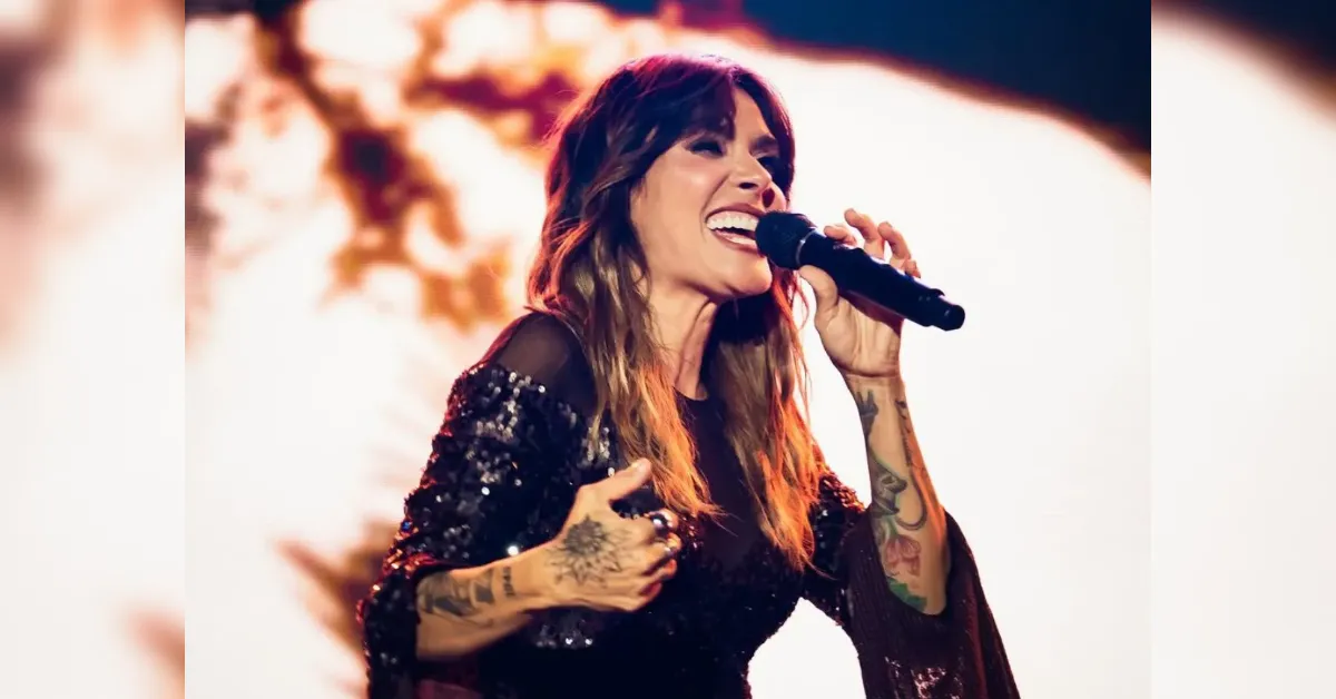 Female singer performing on stage, holding a microphone and singing passionately, wearing a sequined outfit with visible tattoos on her arms.