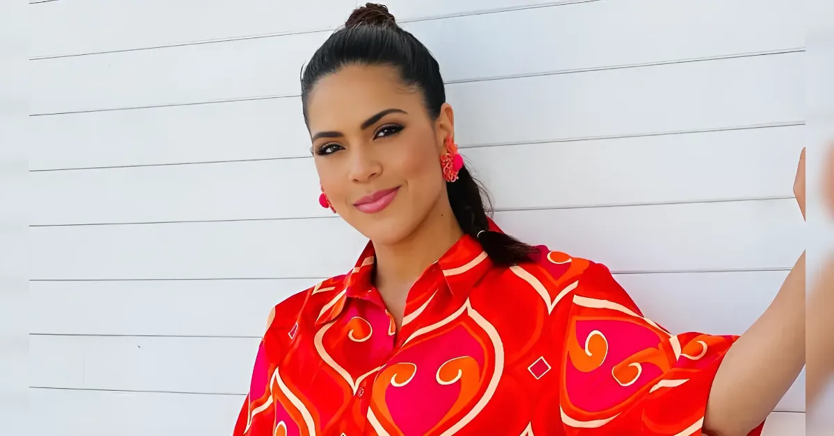 Woman with dark hair in a ponytail wearing a red patterned shirt, smiling against a white paneled wall.