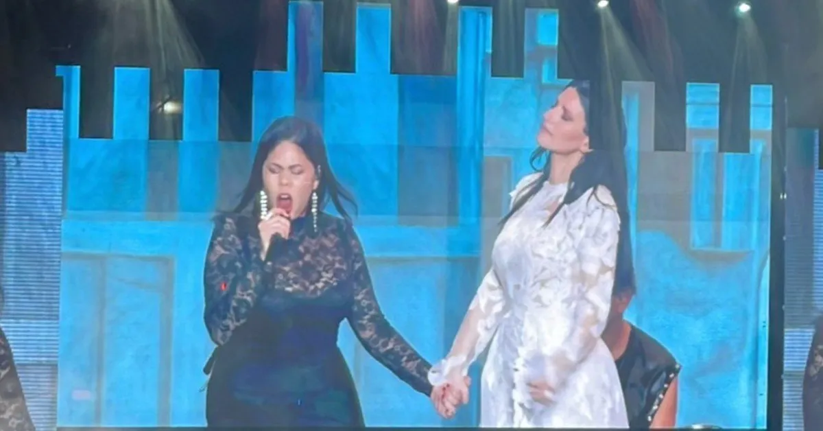 Two female singers on stage holding hands; one in a black lace dress singing into a microphone, the other in a white dress with eyes closed.