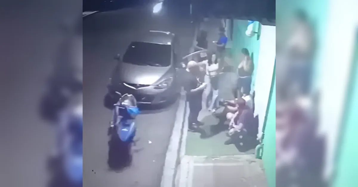 Nighttime street scene with a silver car and blue motorcycle parked by the curb as several people stand or sit along a green storefront.