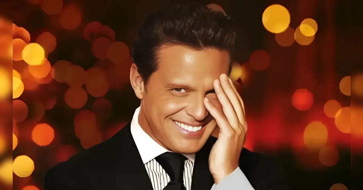 Smiling man in a black suit and white shirt, with his hand partially covering his face, against warm orange bokeh lights.