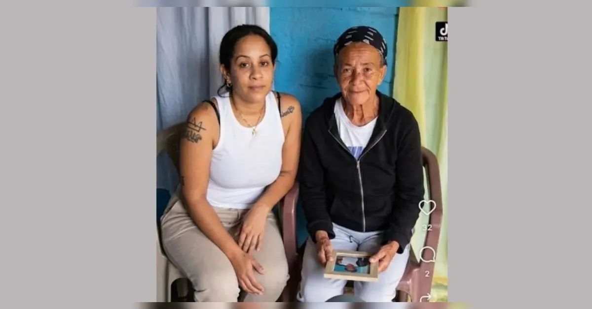 Two women sit together; younger woman with tattoos in a white tank top beside an elderly woman holding a photo on a tablet.