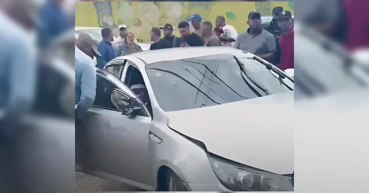 Crowd of people surrounding a silver sedan with a damaged front end after a collision. The driver's door is open and onlookers inspect the scene.