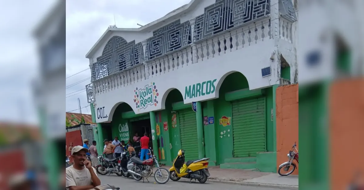 Green storefront with arched entryways and signs for Kola Real and MARCOS; people gathered outside and motorcycles parked along the curb.