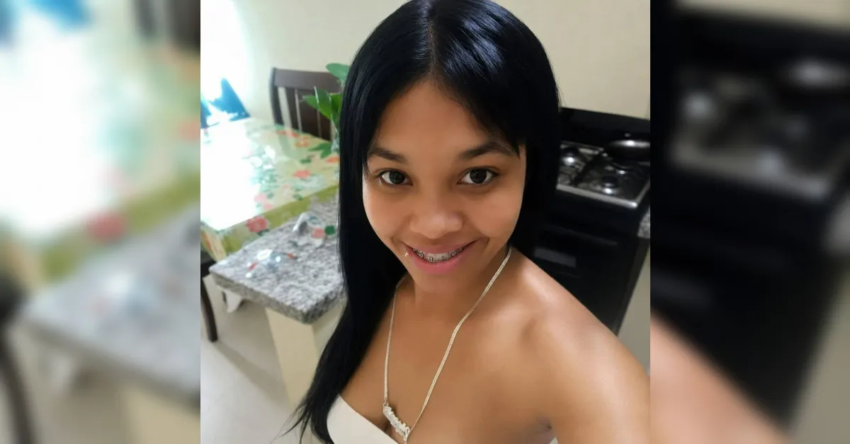 Young woman with long black hair smiling at the camera in a kitchen; braces visible; wearing a white top and necklace.