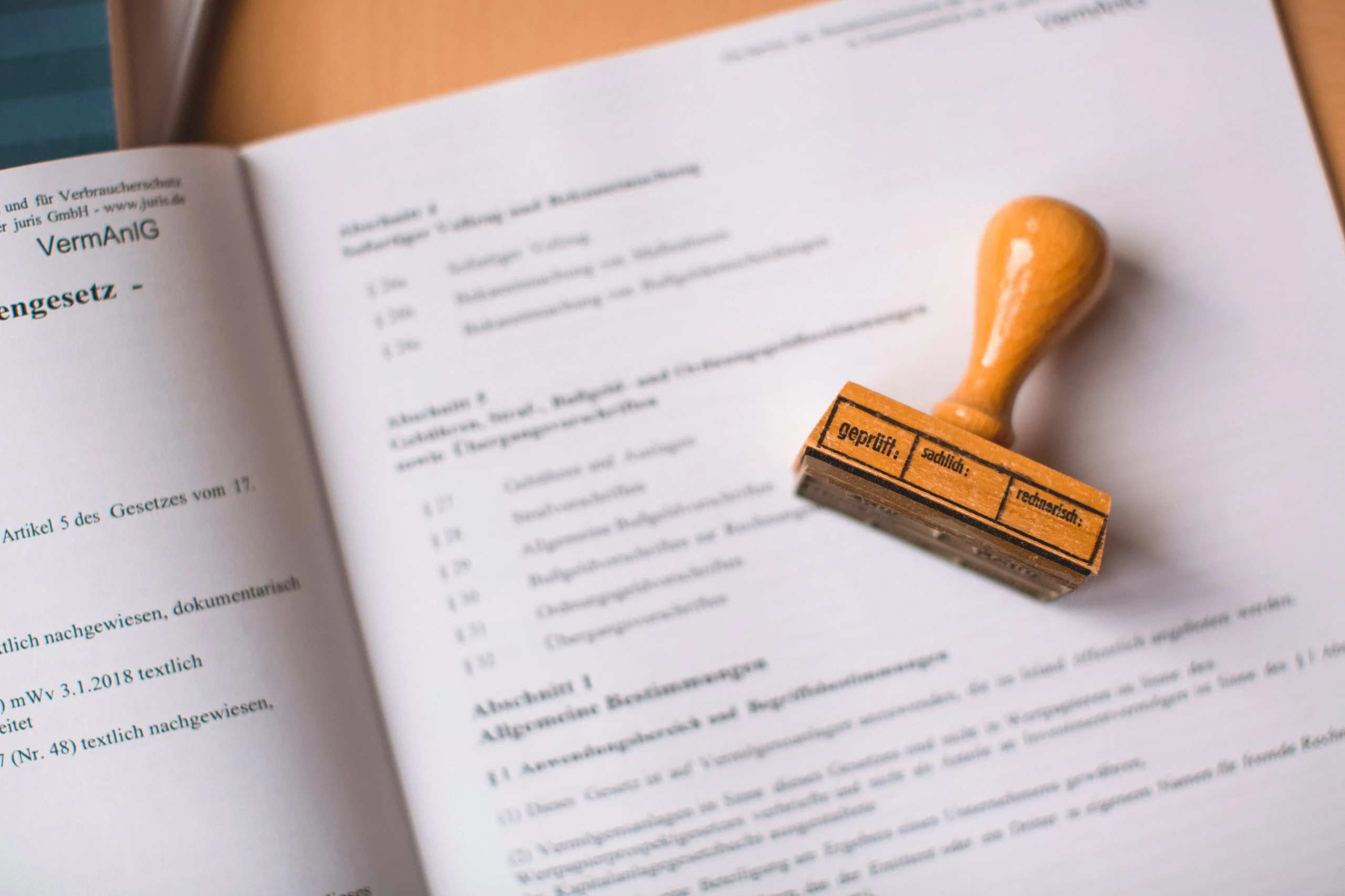 Open book with a wooden stamp resting on the page, stamp labeled with 'geprüft' and related fields in German, resting diagonally across the text.