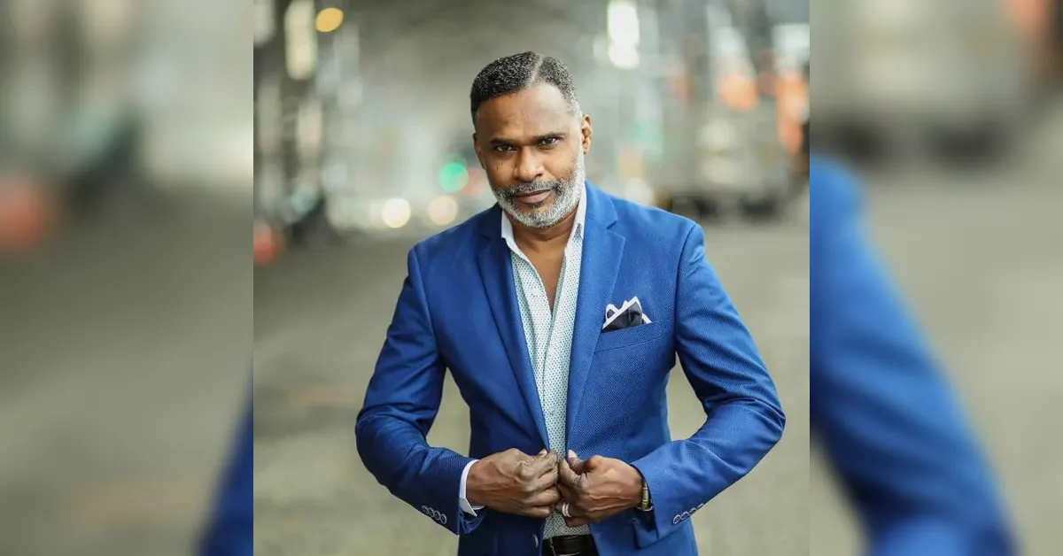 Man in a sharp blue suit adjusts his jacket in a city street backdrop, looking confident at the camera.