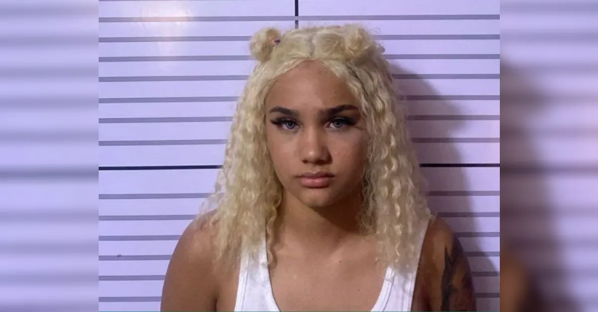 Mugshot of a young woman with platinum blonde curly hair in a white tank top, standing against a height chart background in a police photo.